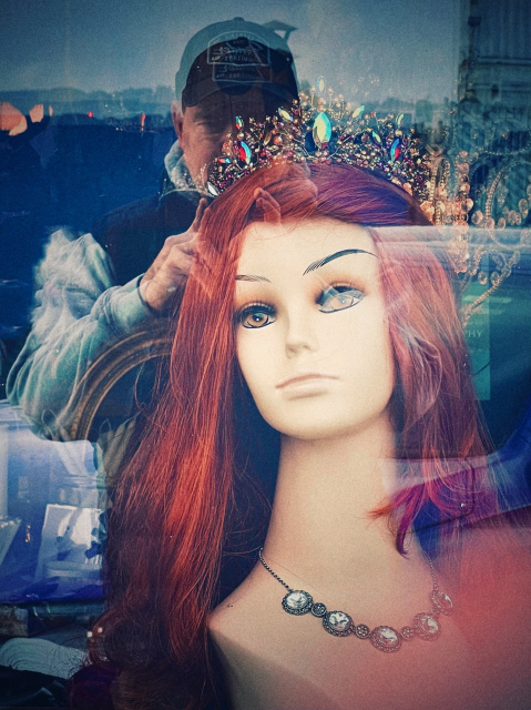 Distorted picture of a mannequin head with long red hair and vintage necklace. Behind is a reflection of the photographer wearing a ball cap.